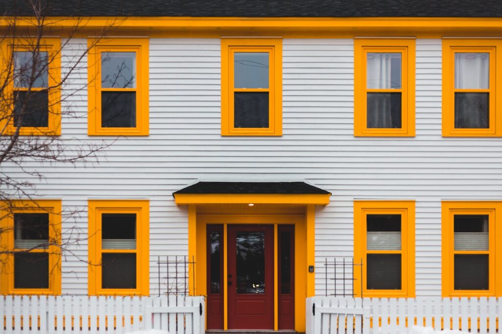 A vibrant yellow-trimmed house with a red door, fence, and multiple windows.